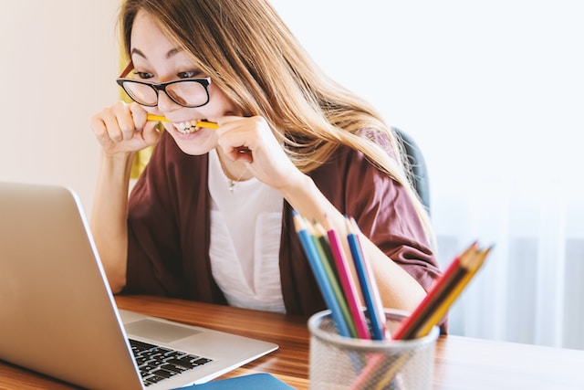 girl with glasses looking at laptop and biting yellow pencil