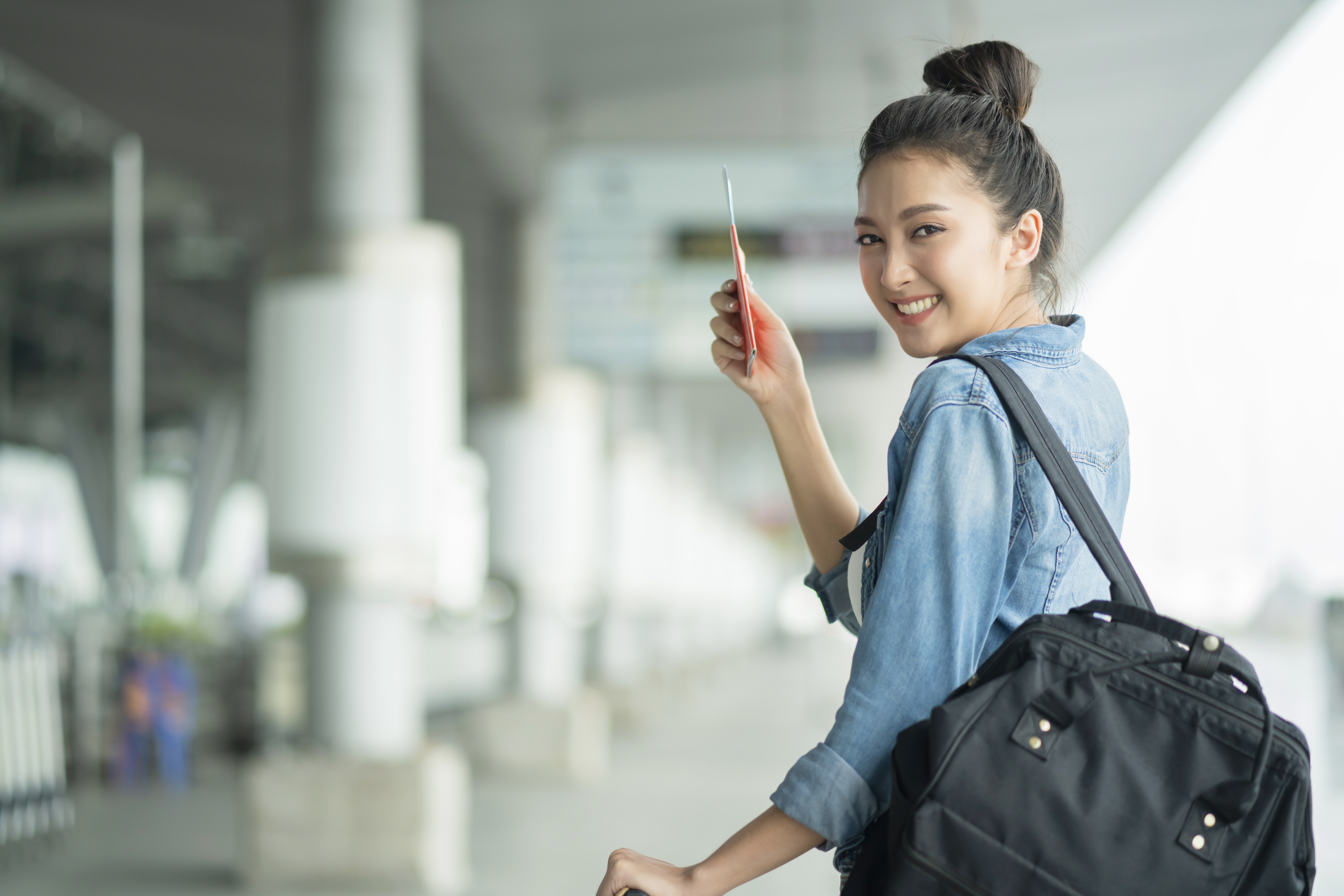 Girl in jean jacket wearing black backpack holding passport at airport traveling 