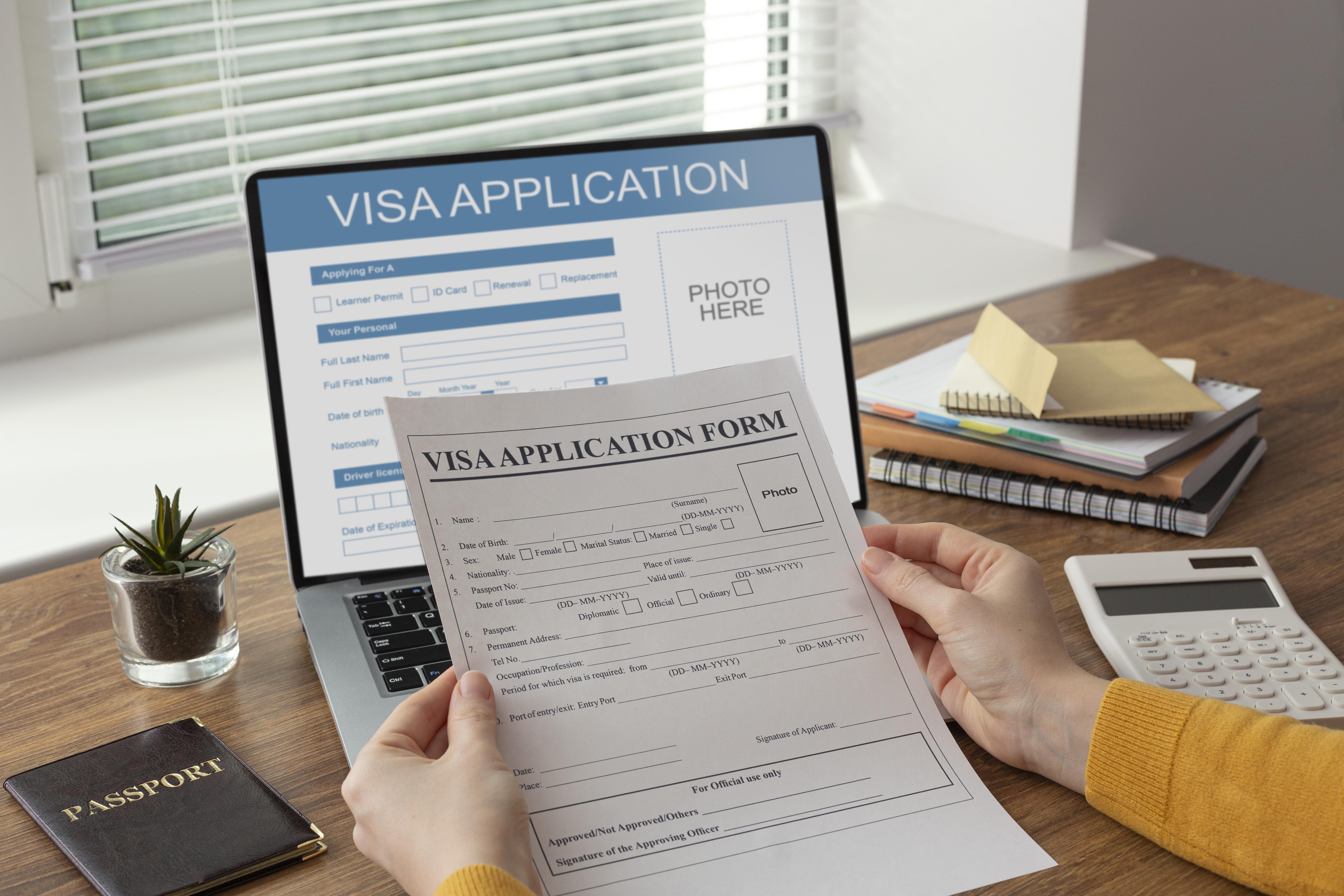 Applying visa on laptop, wooden table with notebooks, calculator, plant and passport, holding visa application form 